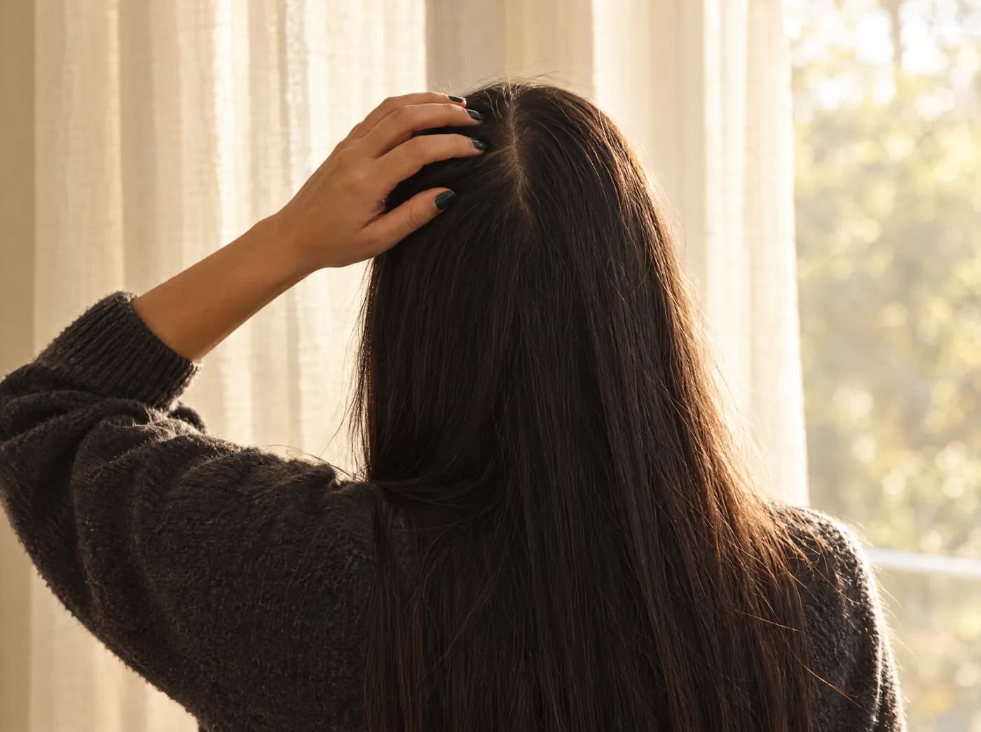 Mujer de espaldas pasándose la mano por un cabello oscuro y largo junto a una ventana con luz cálida